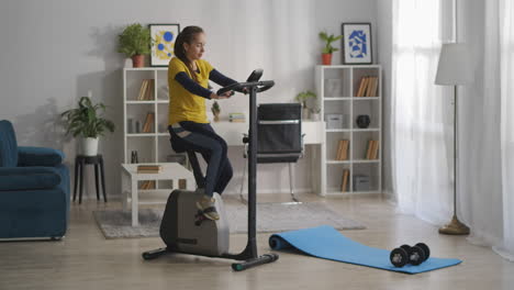 full-length-shot-of-living-room-with-sport-equipment-young-woman-is-training-on-exercise-bike-workout-and-healthy-lifestyle-home-fitness